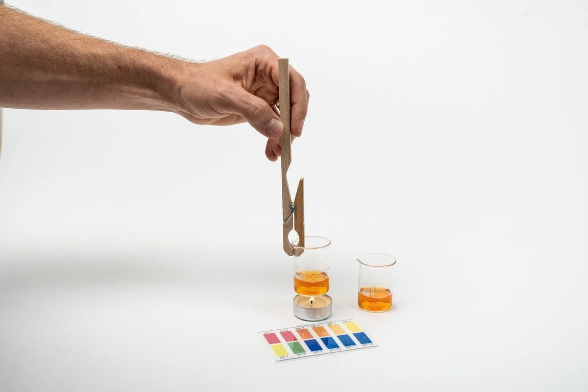 Foto: zwei kleine Gläser mit orangener Flüssigkeit, eines steht, das andere wird von einer Hand die eine Klammer hält über ein Teelicht gehalten. Vor dem Teelicht liegt eine farbige Indikationskarte für verschiedene PH-Werte