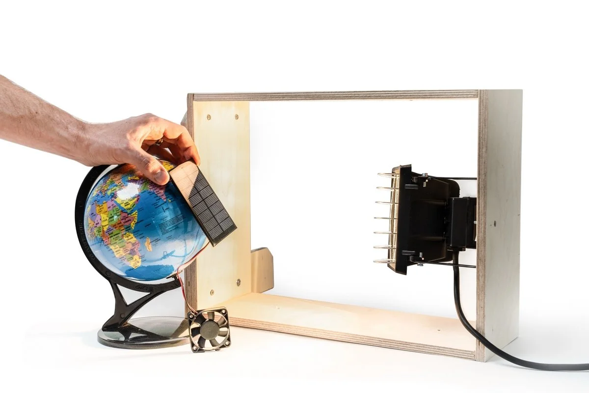 Foto: eine Hand hält ein kleines Solarpanel vor einen kleinen Globus. Angeschlossen ist ein kleiner Propeller. Auf das Solarpanel ist eine Lampe gerichtet, die in einem Holzrahmen befestigt ist.