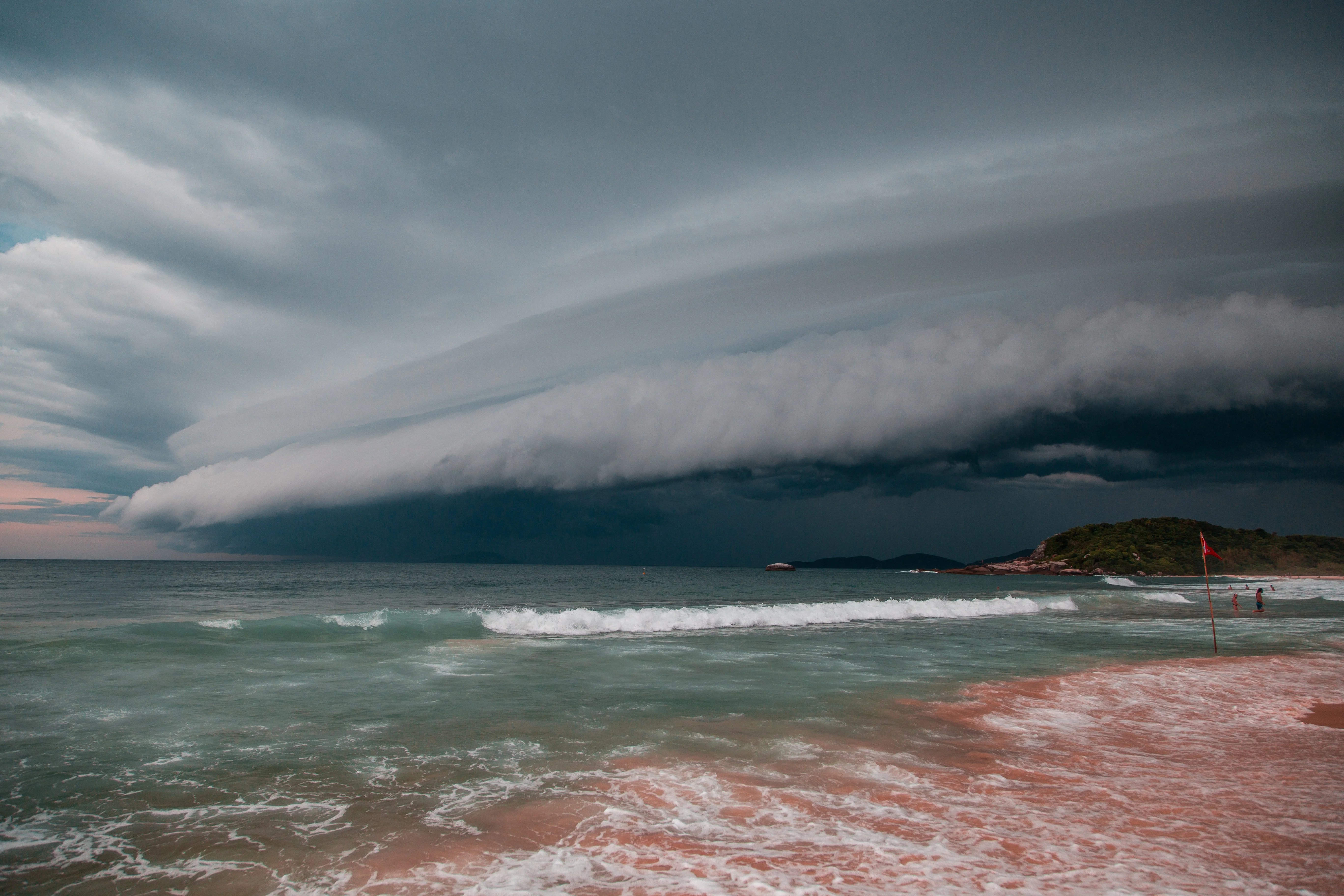 Photo: Beach with sea in the foreground, gusty winds in the background. A flag on the beach indicates strong winds.